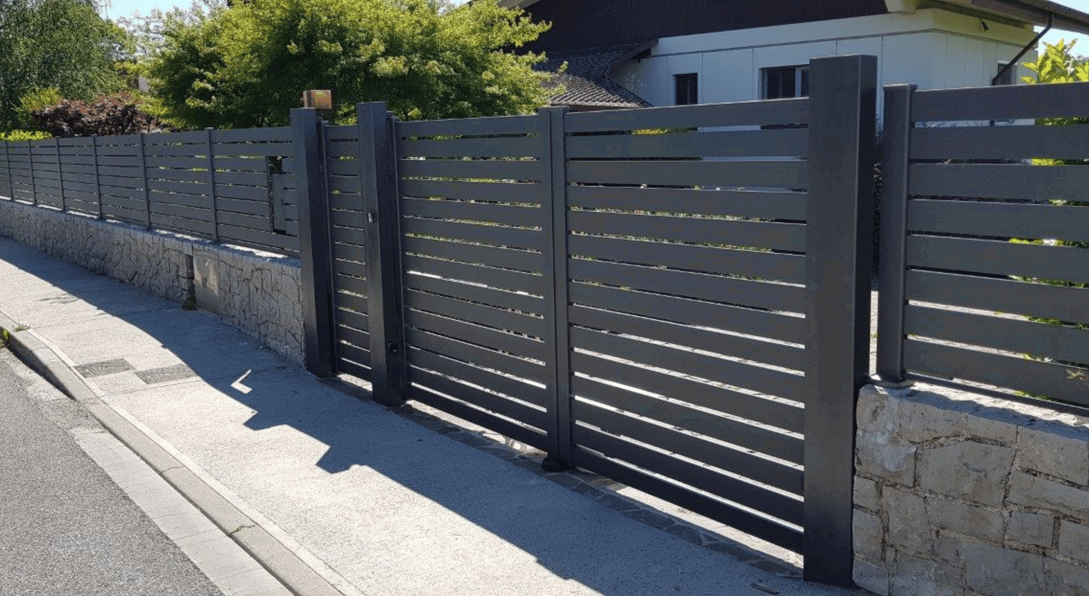 Antracitna aluminijasta dvoriščna ograja in enokrilna vrata z vodoravnimi lamelami na kamnitem zidu.