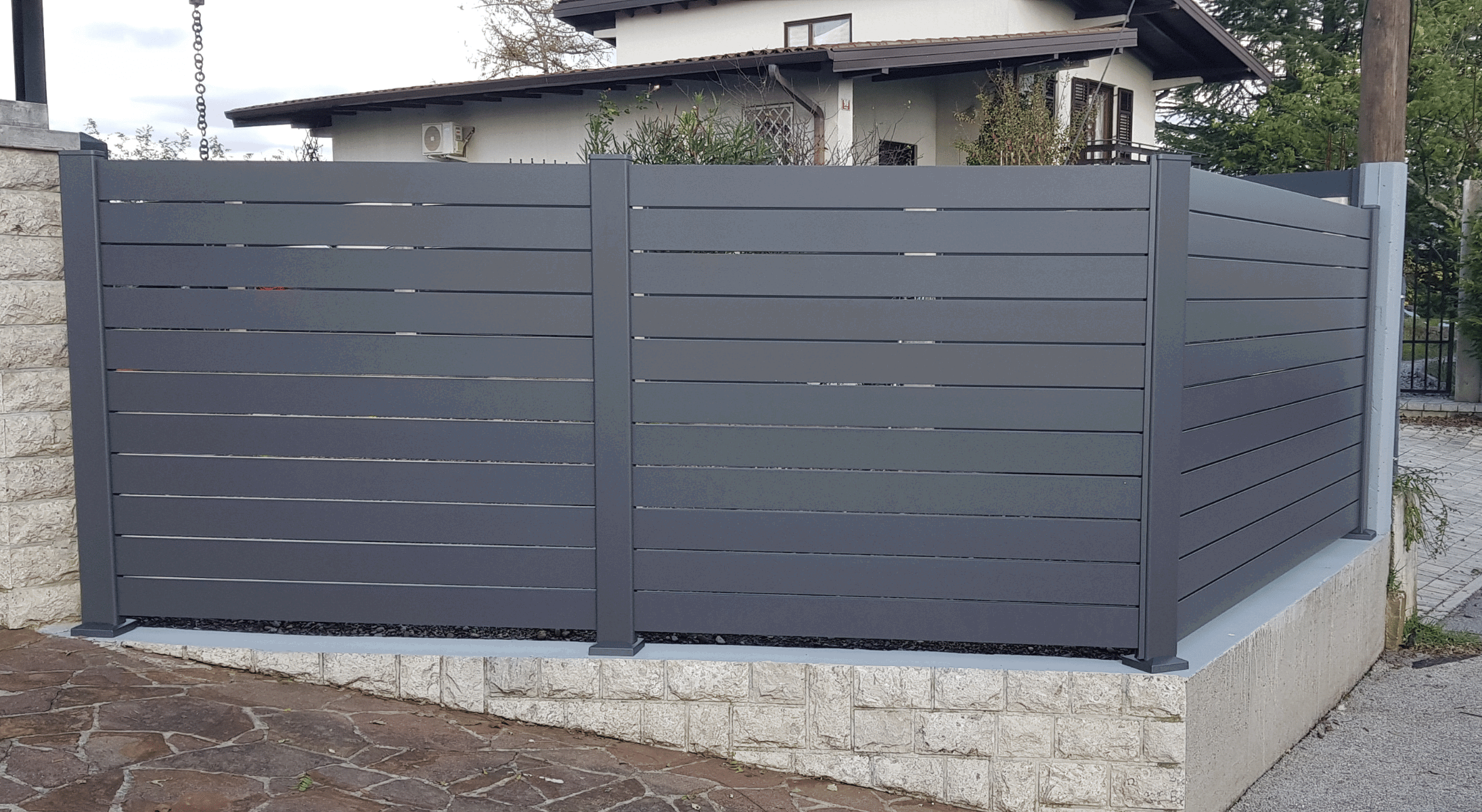 Antracitna aluminijasta dvoriščna ograja z gosto razporejenimi vodoravnimi lamelami na kamnitem podstavku.