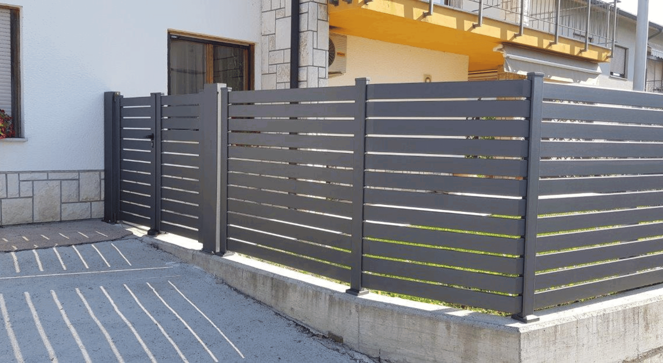 Antracitna aluminijasta dvoriščna ograja z vodoravnimi lamelami ob stanovanjski hiši.